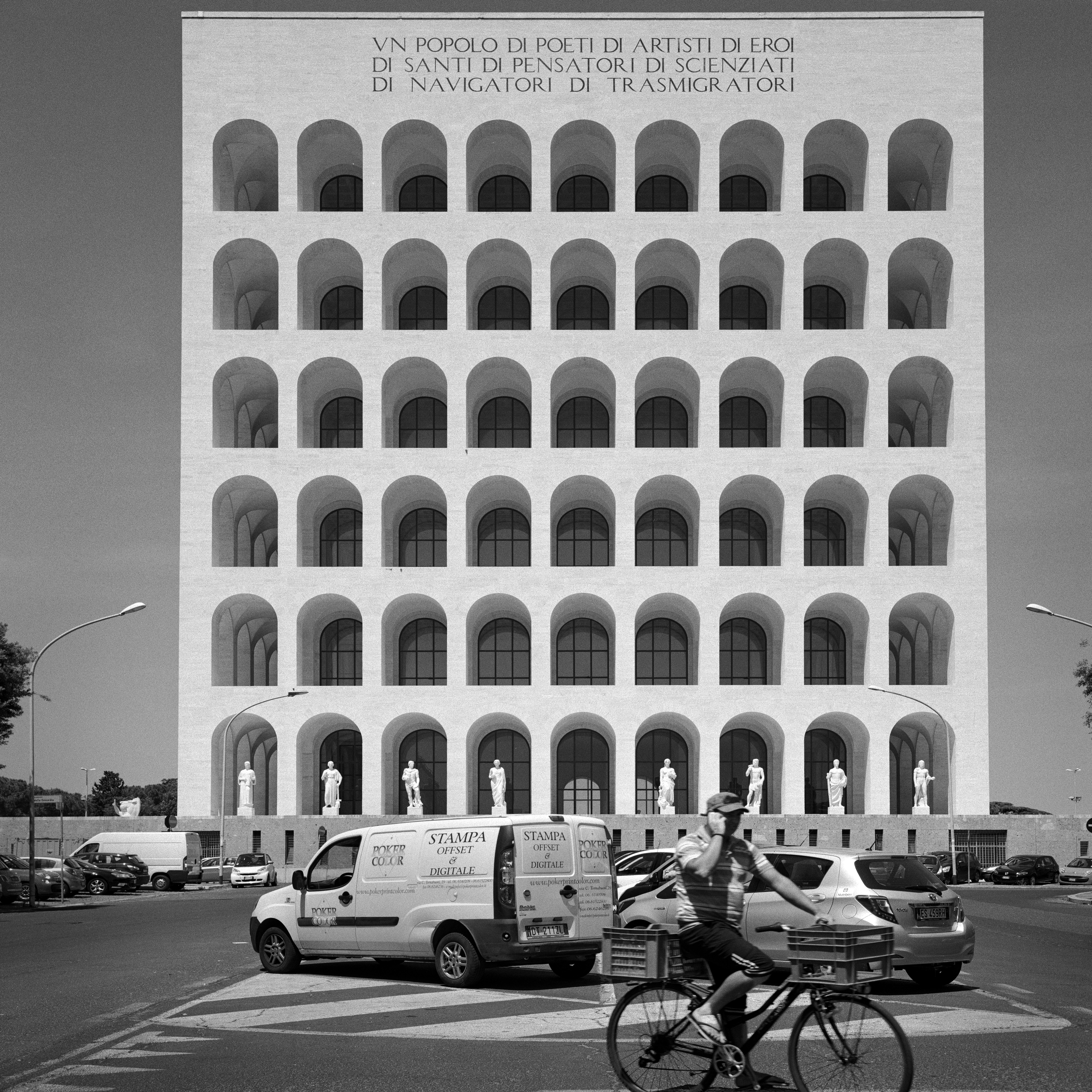 black and white image of a building in Rome with arched windows. A car park in the foreground, a man on a bicycle appears to be talking on the phone